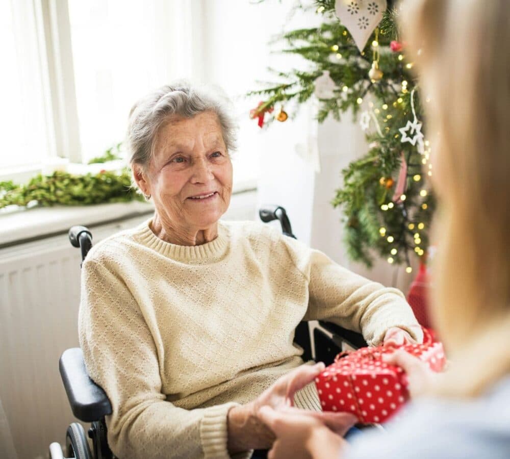 An older female adult with grey hair sitting on a wheelchair beside a Christmas tree while receiving a gift