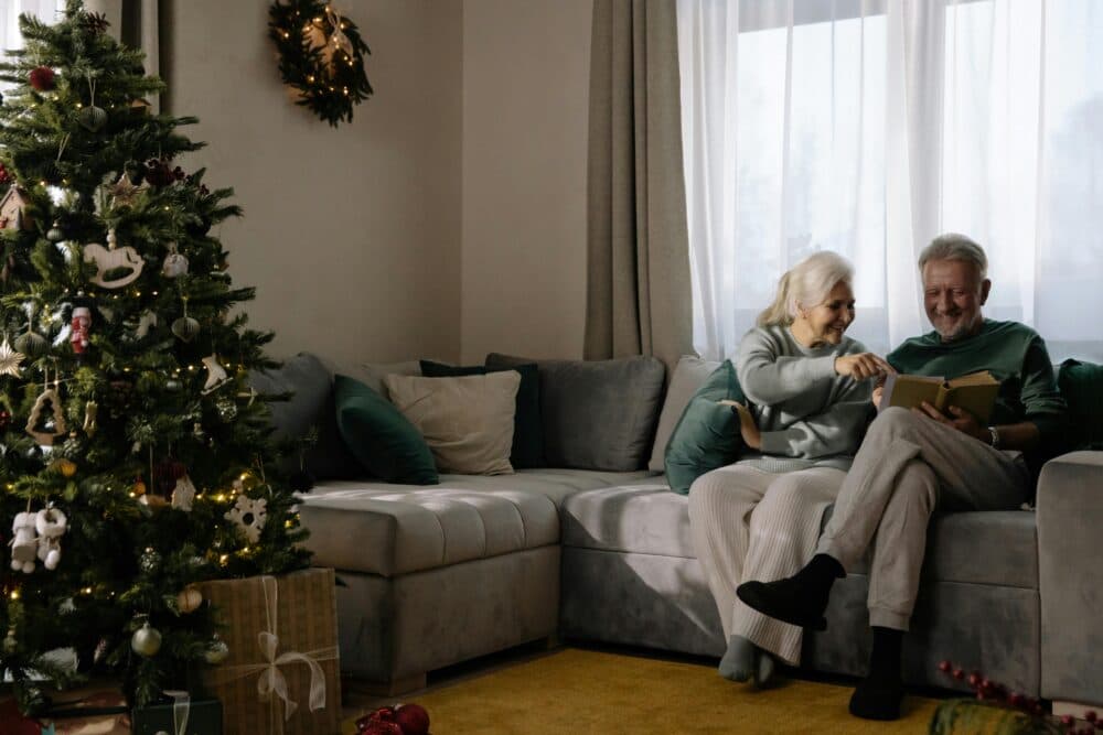 An elderly couple smiles while sitting on a sofa and looking at a book near a decorated Christmas tree. - Home Instead