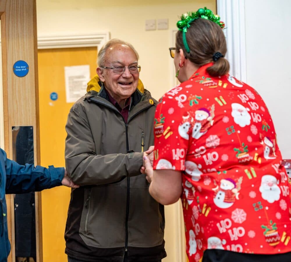 A woman wearing Christmas short and hat greeting an older male adult happy and smiling while wearing eyeglasses and winter clothes while entering the door