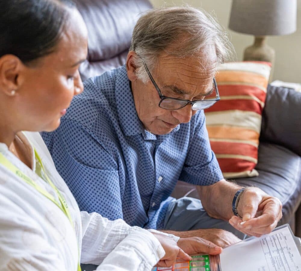 An older male adult with grey hair and wearing eyeglasses taking his medicine while being helped by her female carer