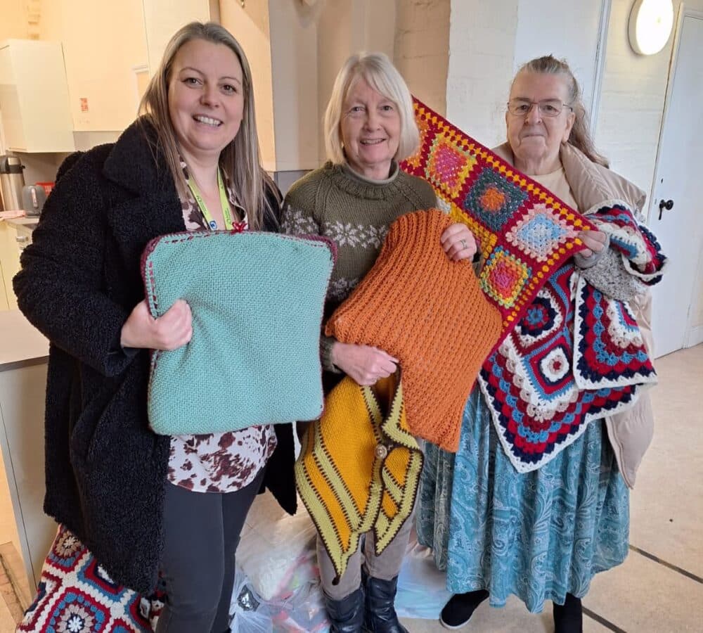 Three women smiling, holding knitted and crocheted blankets and cushions, standing indoors. - Home Instead