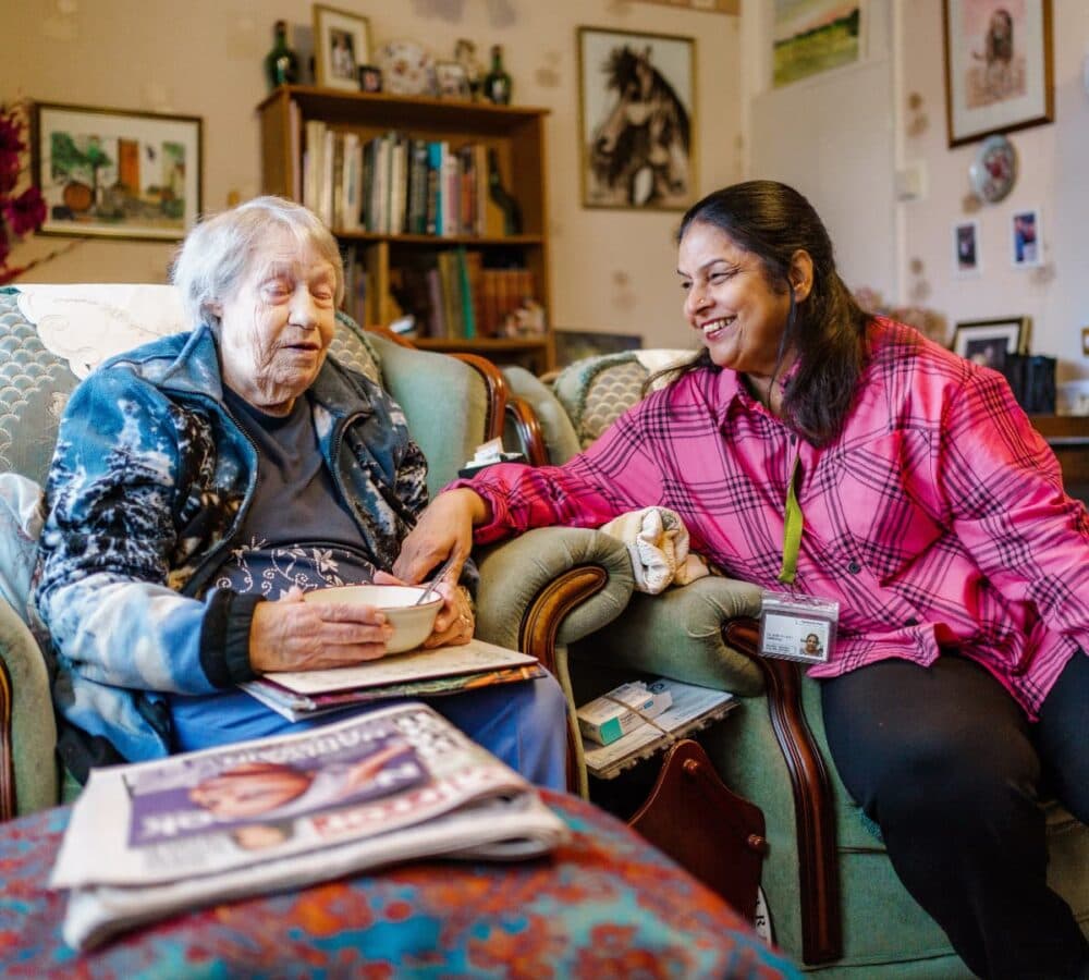 Two women happy and smiling while chatting and sitting on the couch inside the house