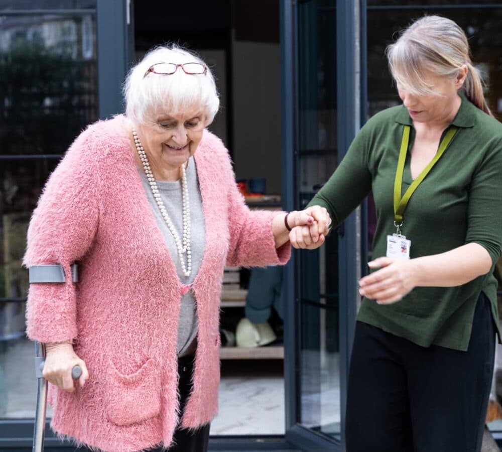 Elderly woman with crutch being assisted by a caregiver outside a building. - Home Instead