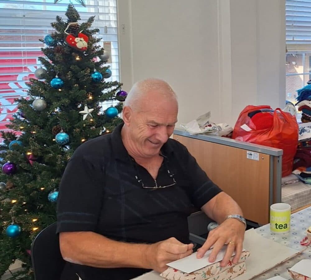 An older man wrapping a gidt at the table with a Christmas tree at his back