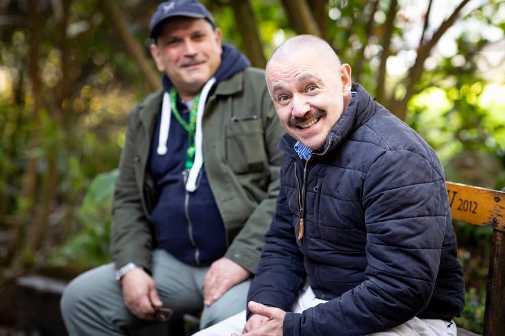 Two men sit outdoors on a bench, smiling warmly with greenery in the background. - Home Instead