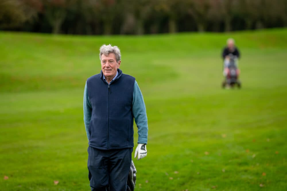 Older man in a blue waistcoat walking on a golf course, another person blurred in the background. - Home Instead