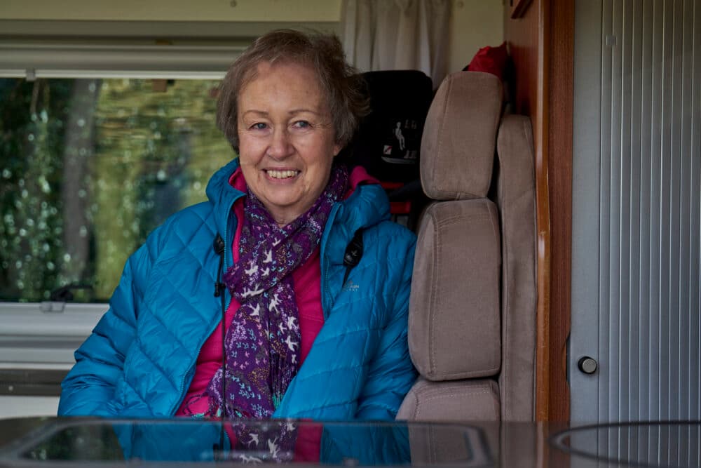 An older woman in a blue jacket smiles whilst sitting inside a camper van. - Home Instead