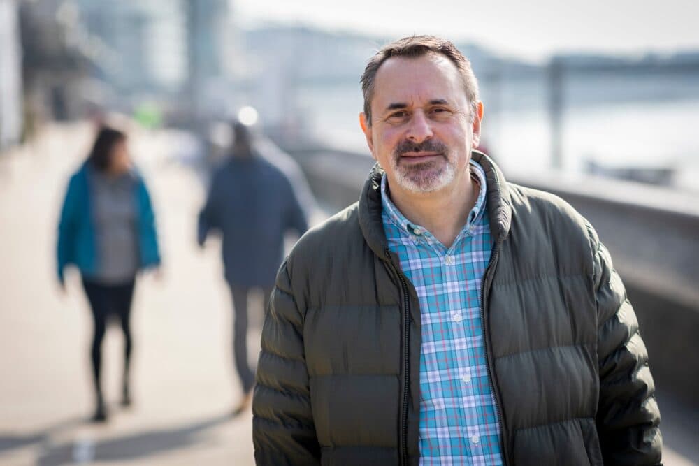 Man in a green jacket smiling outdoors on a sunny urban walkway, people blurred in the background. - Home Instead