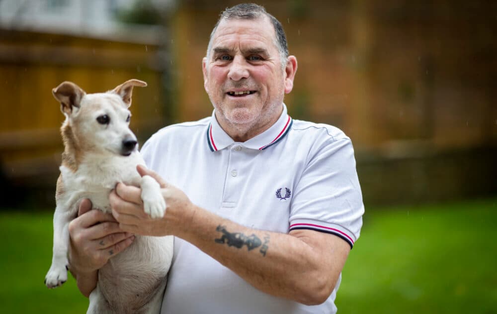 Smiling man in a white polo shirt holding a small brown and white dog outside in a garden. - Home Instead