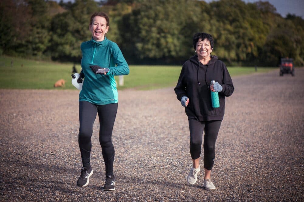 Two women in sportswear jog outdoors on a gravel path, smiling and holding water bottles. - Home Instead
