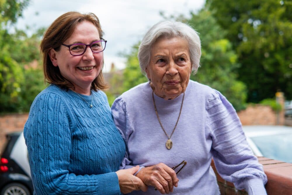 Middle-aged woman and elderly woman smiling and standing together outside by a brick wall on a cloudy day. - Home Instead