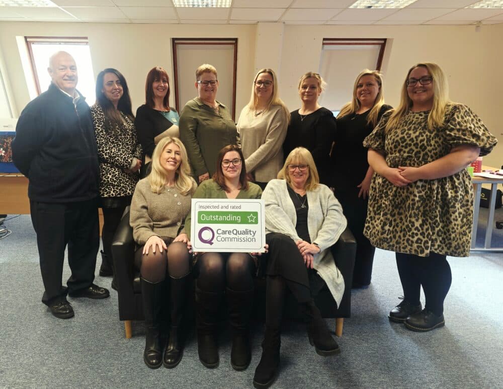 A group of twelve people, mostly women, pose indoors with a Care Quality Commission "Outstanding" sign. - Home Instead