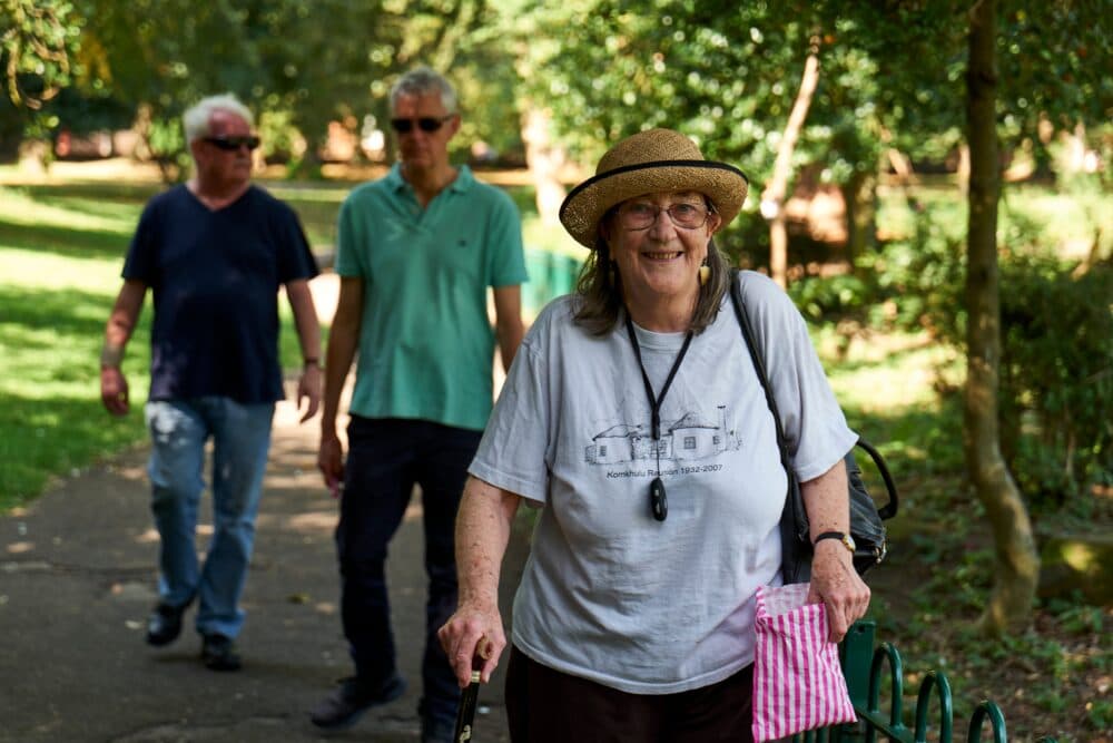 Smiling elderly woman with a walking stick walks in a park; two men are walking behind her on the path. - Home Instead