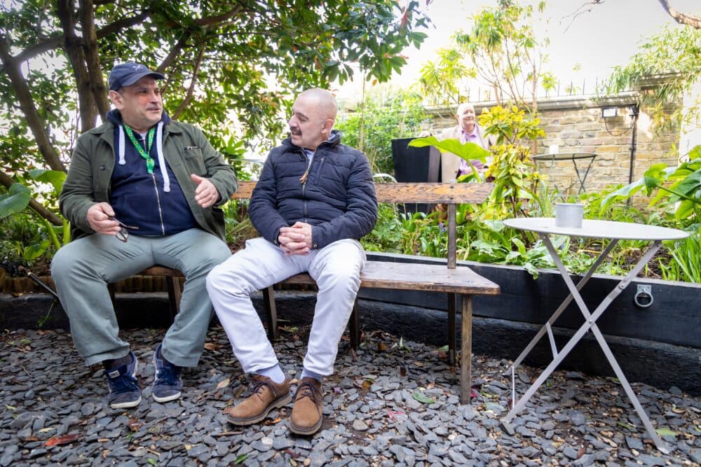 Two men sit and talk on a wooden bench in a leafy garden, with a woman standing in the background. - Home Instead