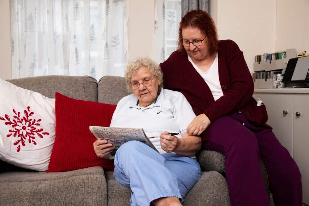 Two women sit on a sofa; one is doing a crossword puzzle while the other sits beside her, smiling supportively. - Home Instead