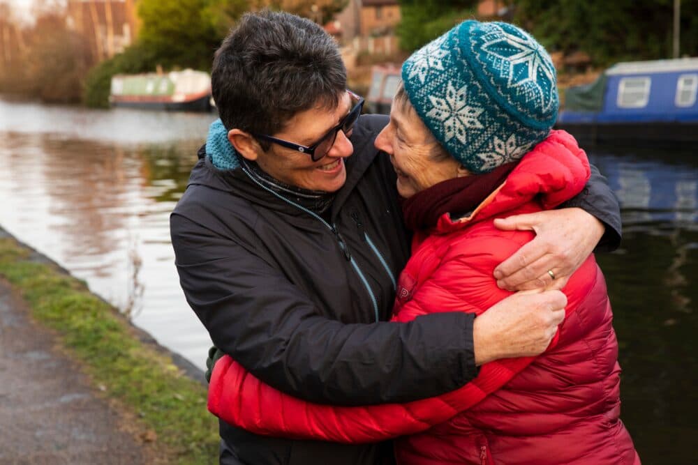 Two people in winter jackets and hats smiling and hugging by a canal with boats in the background. - Home Instead