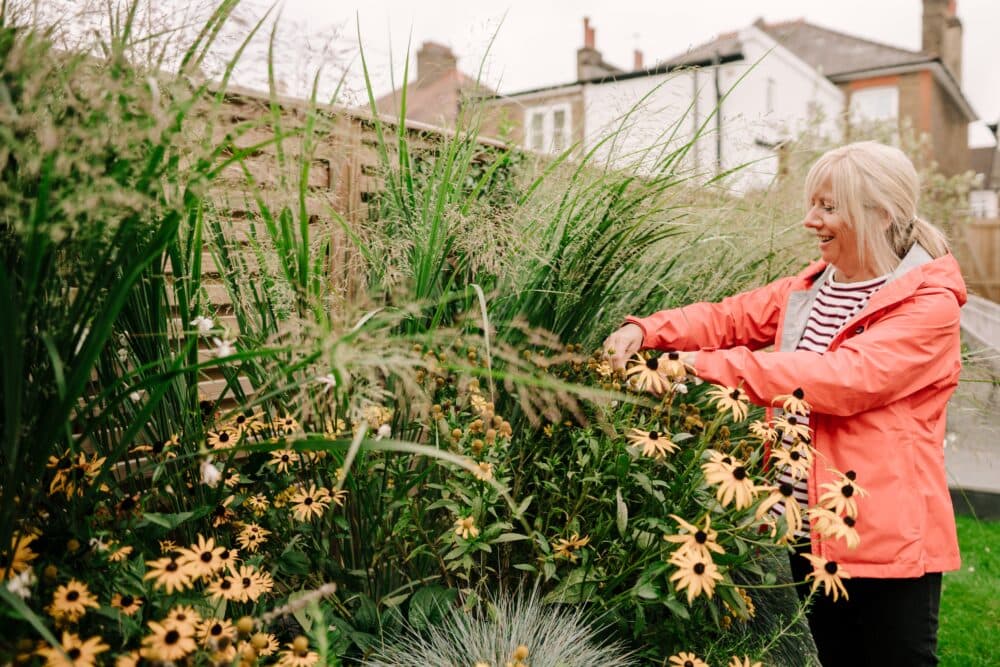 Woman in a coral jacket tending to yellow flowers and tall grass in a garden. - Home Instead
