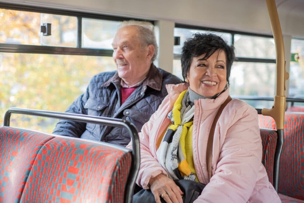 Smiling older couple sitting together on a bus with patterned seats and sunlight coming through the windows. - Home Instead