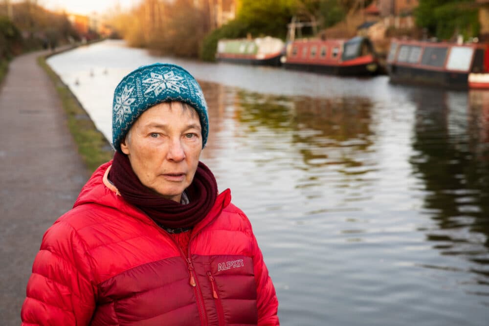 An older woman in a red jacket and blue hat stands by a canal with boats and a path in the background. - Home Instead