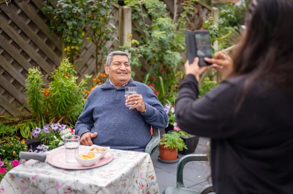 Smiling man holding a glass of water is photographed by a woman in a garden with flowers on the table. - Home Instead
