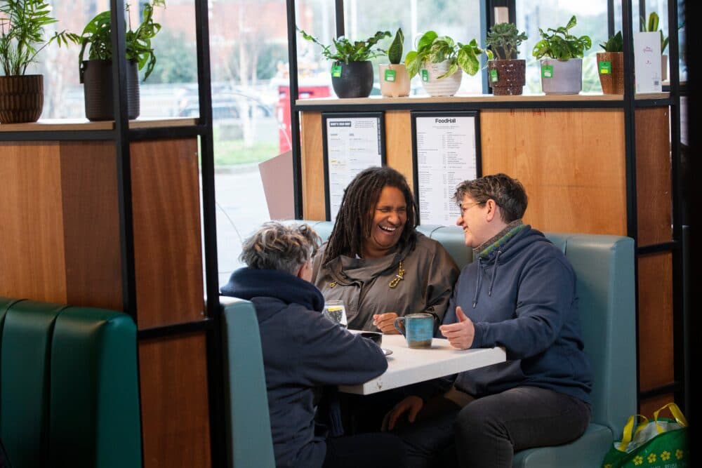 Three people sit at a café booth, smiling and talking over drinks, with plants on the divider behind them. - Home Instead