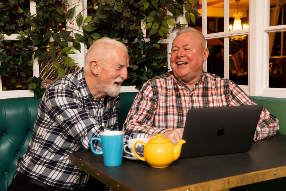Two older men laughing together at a table with a laptop, teapot, and mugs, sitting in a cosy café. - Home Instead