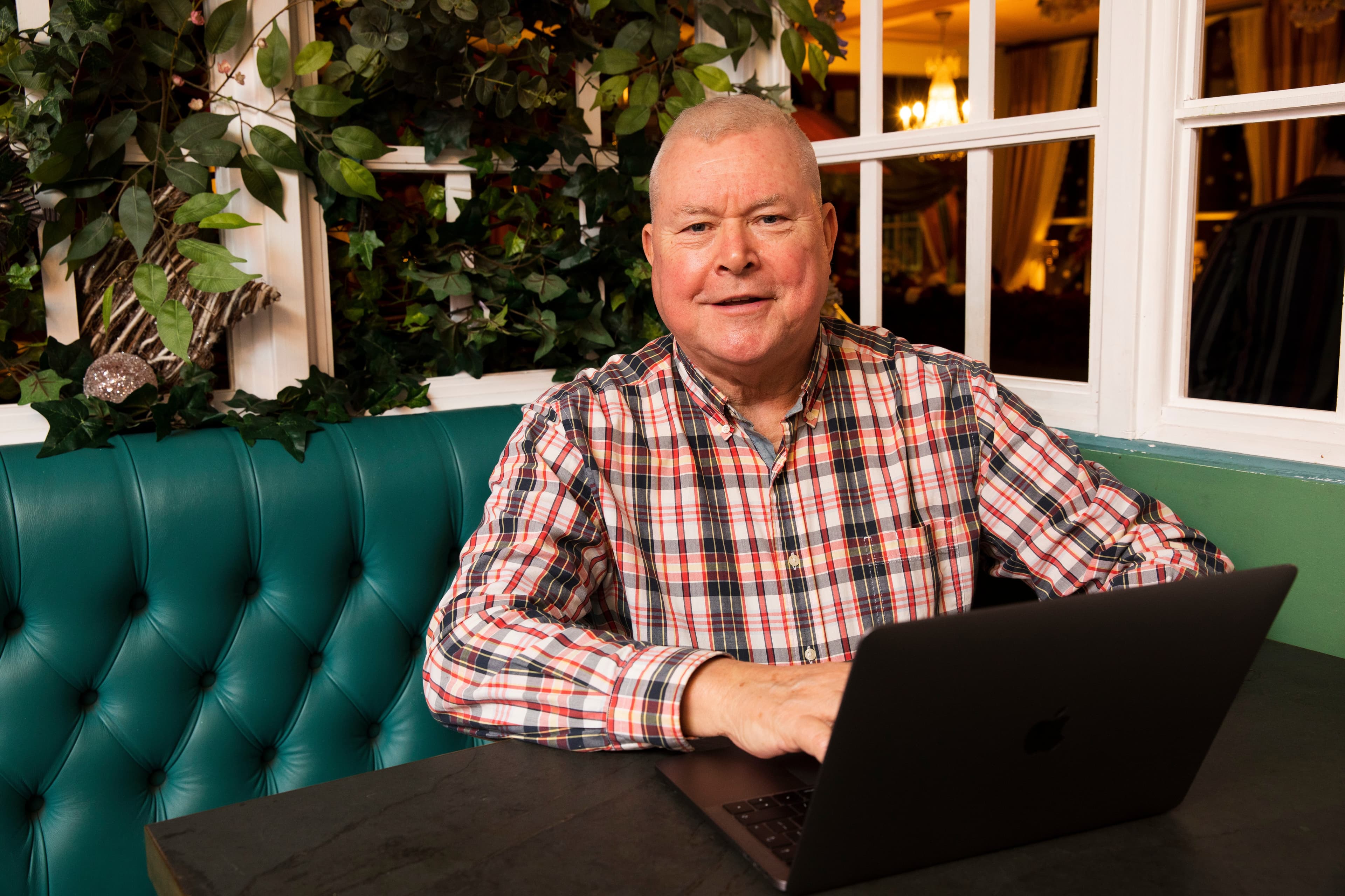 Smiling man in a checked shirt sits at a table with a laptop in a cosy, plant-filled room. - Home Instead