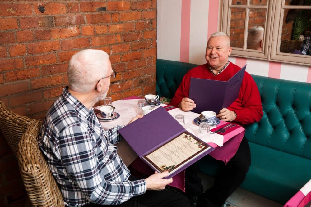 Two older men sit in a café, smiling and looking at menus with teacups on the table. - Home Instead