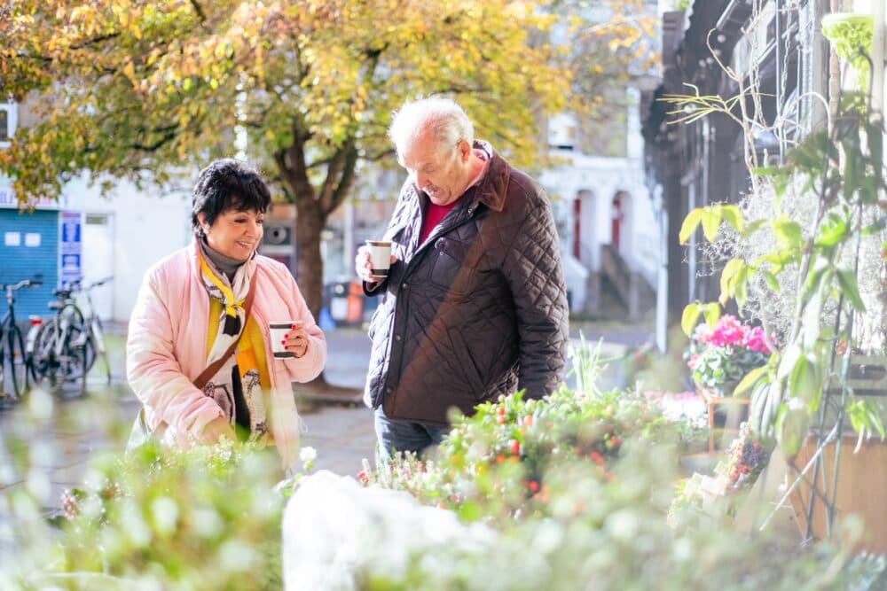 An older couple drink coffee while browsing plants at an outdoor market on a sunny day. - Home Instead