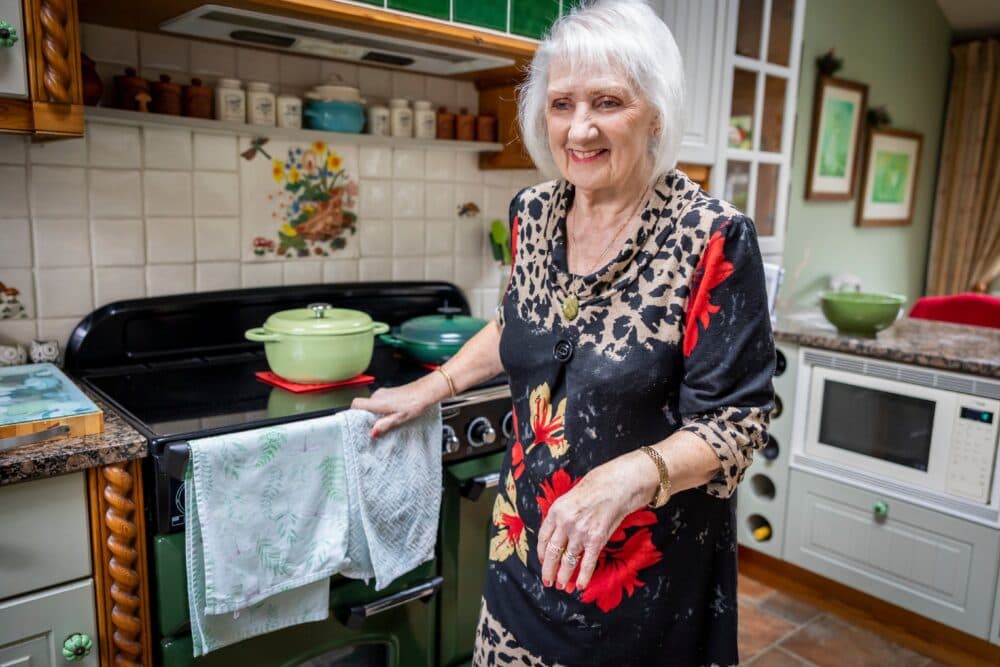 Smiling elderly woman stands by a cooker in a cosy, colourful kitchen with tea towels hanging. - Home Instead