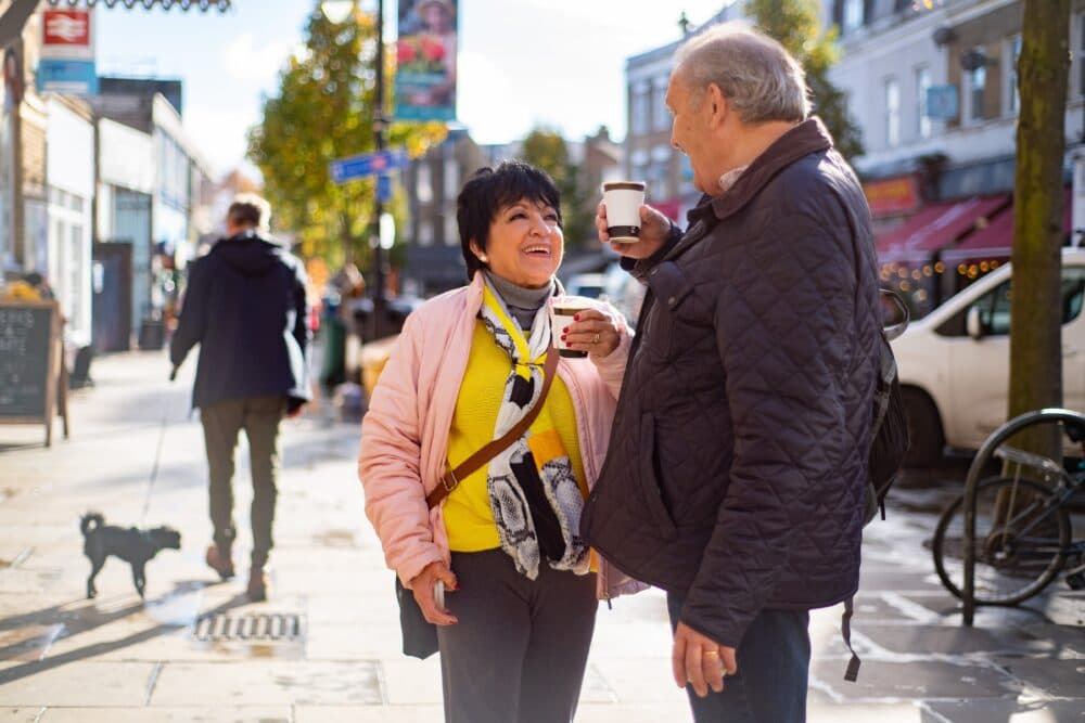 An older couple smile and enjoy coffee together while walking on a sunny city street. - Home Instead