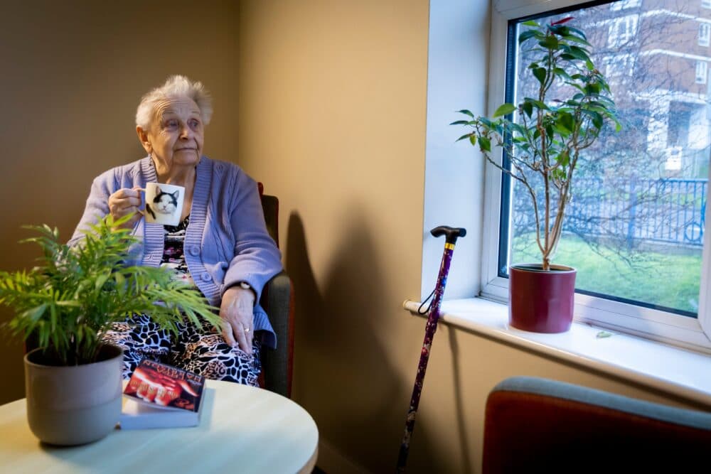 Elderly woman in a chair holds a mug, with books, plants, and a walking stick by the window. - Home Instead