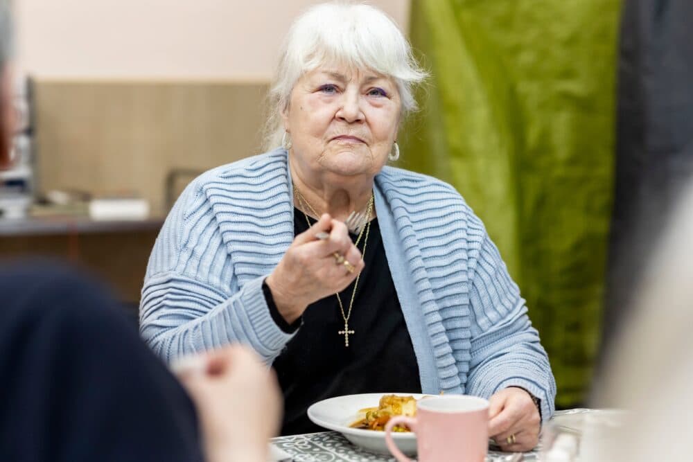 Elderly woman in a blue jumper eating a meal at a table, holding a fork, with a pink mug nearby. - Home Instead