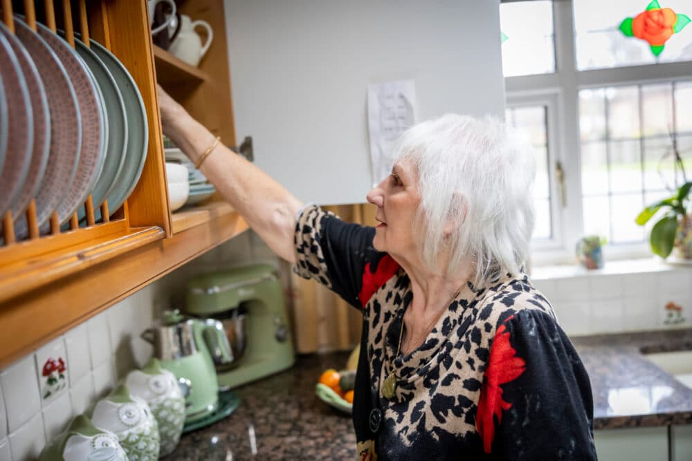 Older woman reaches for a bowl on a kitchen shelf next to a worktop with a mixer and jars. - Home Instead