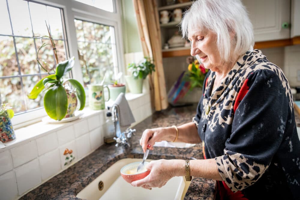 Older woman standing at a kitchen sink, stirring food in a bowl, with plants on the windowsill nearby. - Home Instead