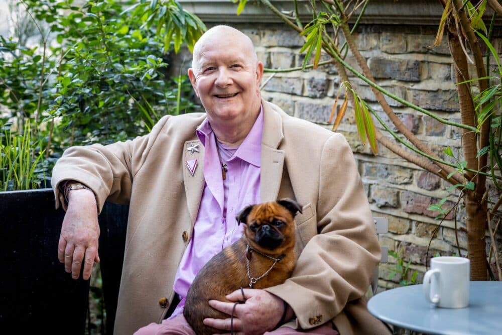 Smiling person in a tan coat holding a small brown dog, sitting outside at a table with a white mug. - Home Instead