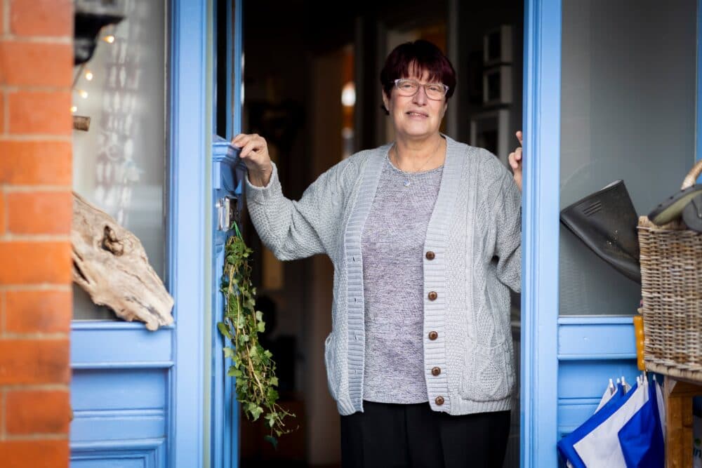 Older woman with short hair and glasses stands smiling in the doorway of a blue-painted home. - Home Instead