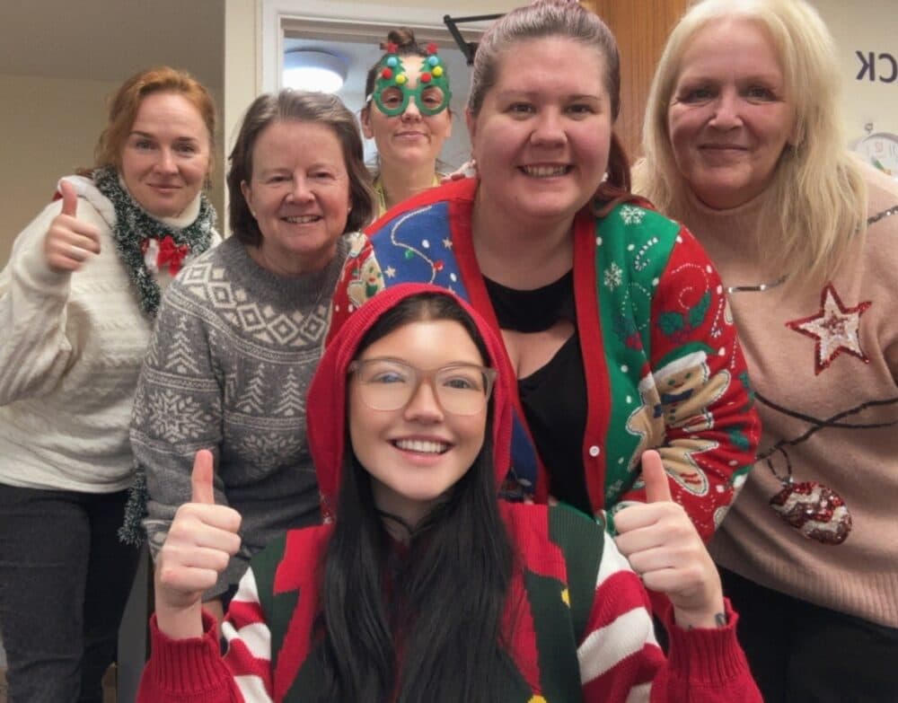 Six smiling women in festive holiday jumpers pose together, some giving thumbs up, in a cheerful indoor setting. - Home Instead