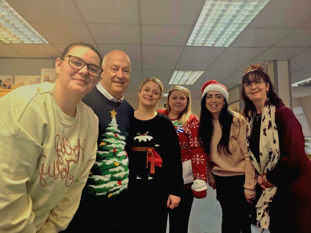 Six people in festive jumpers and hats smiling together in an office setting. - Home Instead