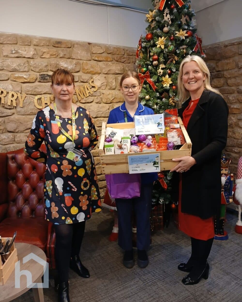 Three women stand by a Christmas tree, holding festive gift hampers and smiling for the camera. - Home Instead