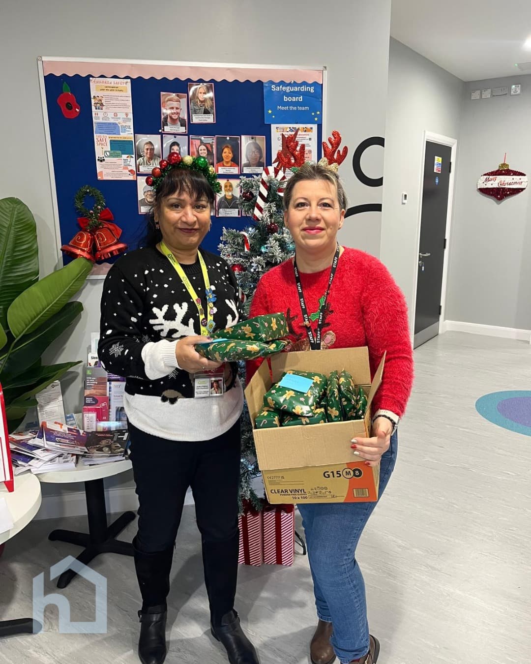 Two women in festive jumpers hold wrapped presents by a Christmas tree in an office setting. - Home Instead