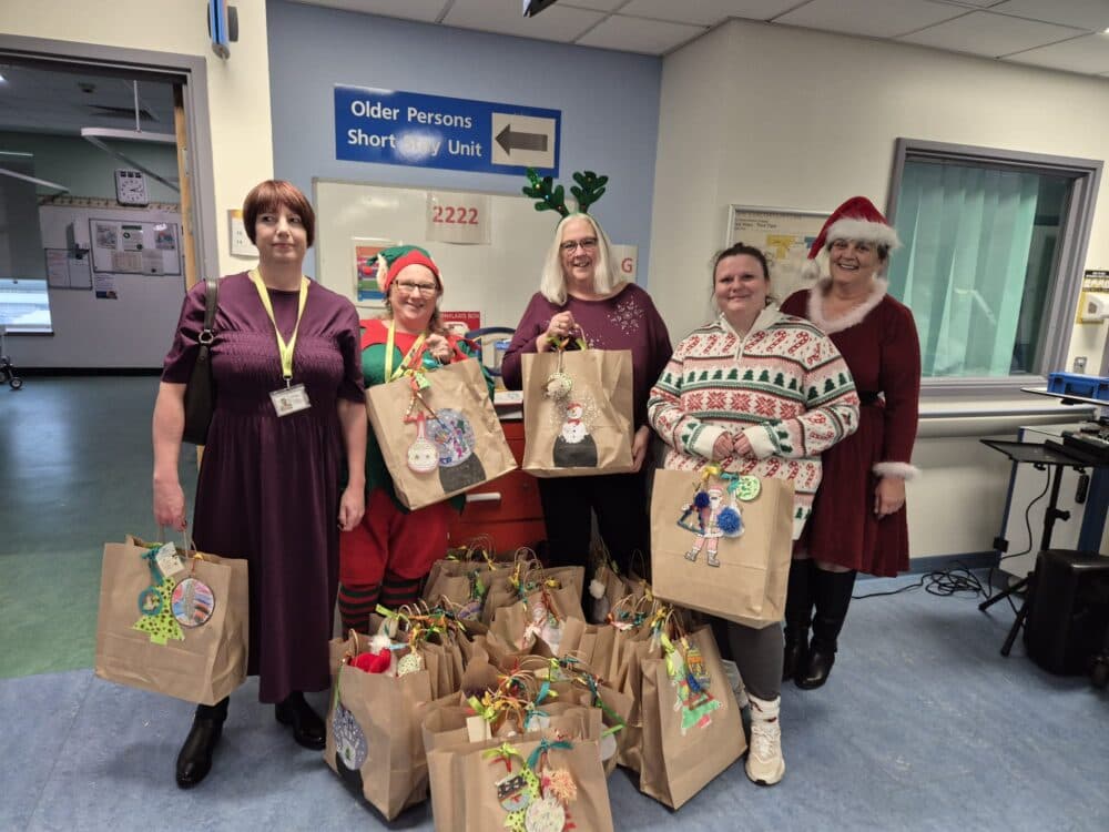 Five women in festive outfits hold decorated gift bags in a hospital corridor, surrounded by more gift bags. - Home Instead