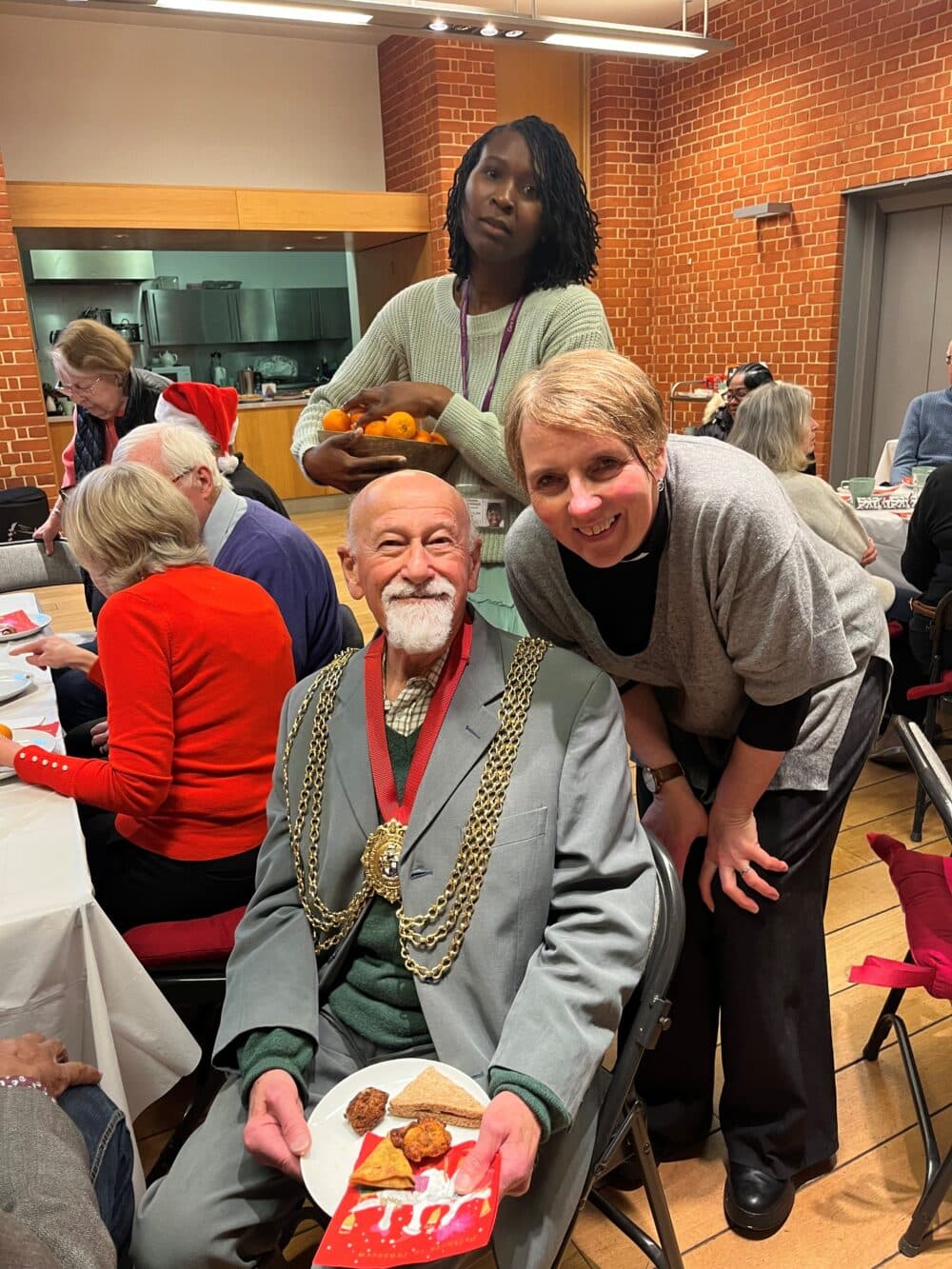 A smiling man in formal attire sits with a plate of food at a festive gathering, surrounded by others. - Home Instead