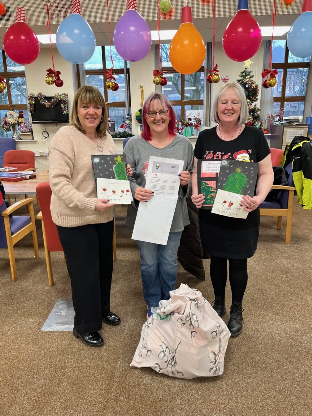 Three women smiling and holding festive cards and a letter, standing in a decorated room with balloons. - Home Instead