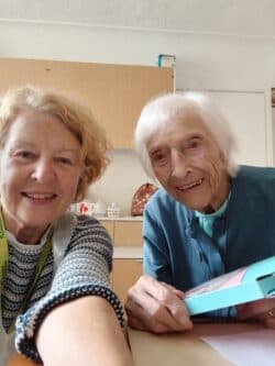 Two elderly women smiling together at a table in a cosy, well-lit room. - Home Instead