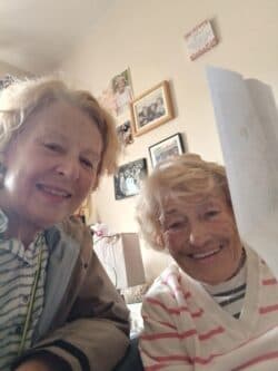 Two elderly women smiling indoors, one holding up a piece of paper, with photos on the wall behind them. - Home Instead