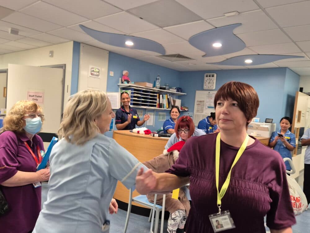 A woman in purple shakes hands with a nurse in a busy hospital reception area with smiling staff. - Home Instead