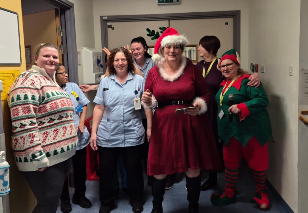 A group of people in festive holiday costumes, including Father Christmas and an elf, pose and smile in a corridor. - Home Instead