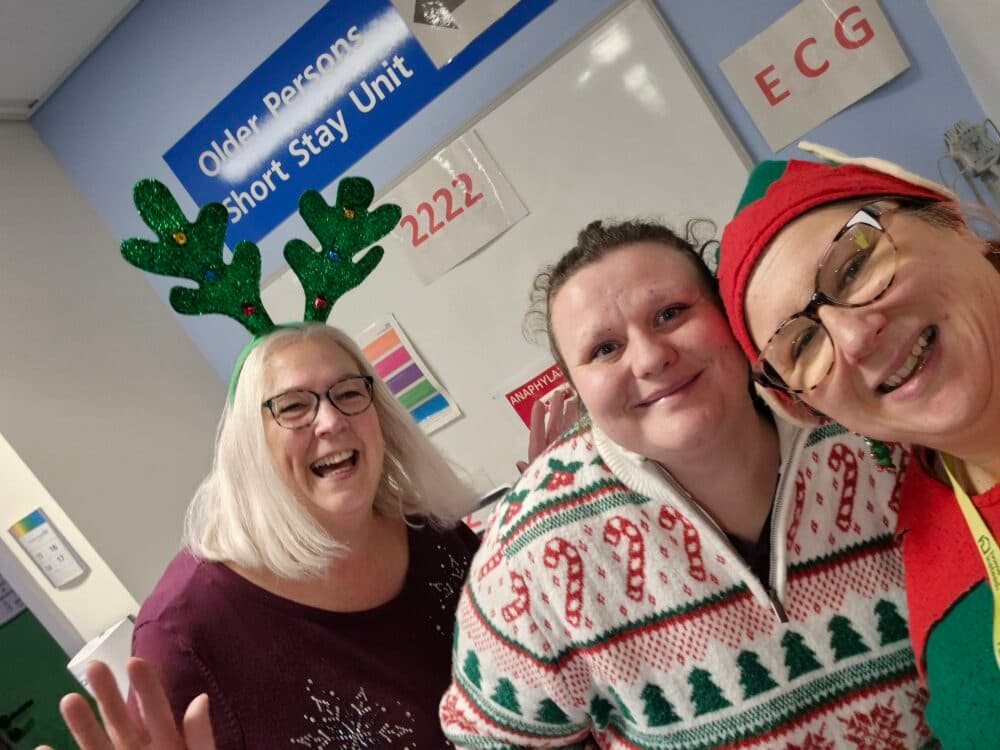 Three smiling adults in festive jumpers and headbands pose in a hospital ward decorated for Christmas. - Home Instead