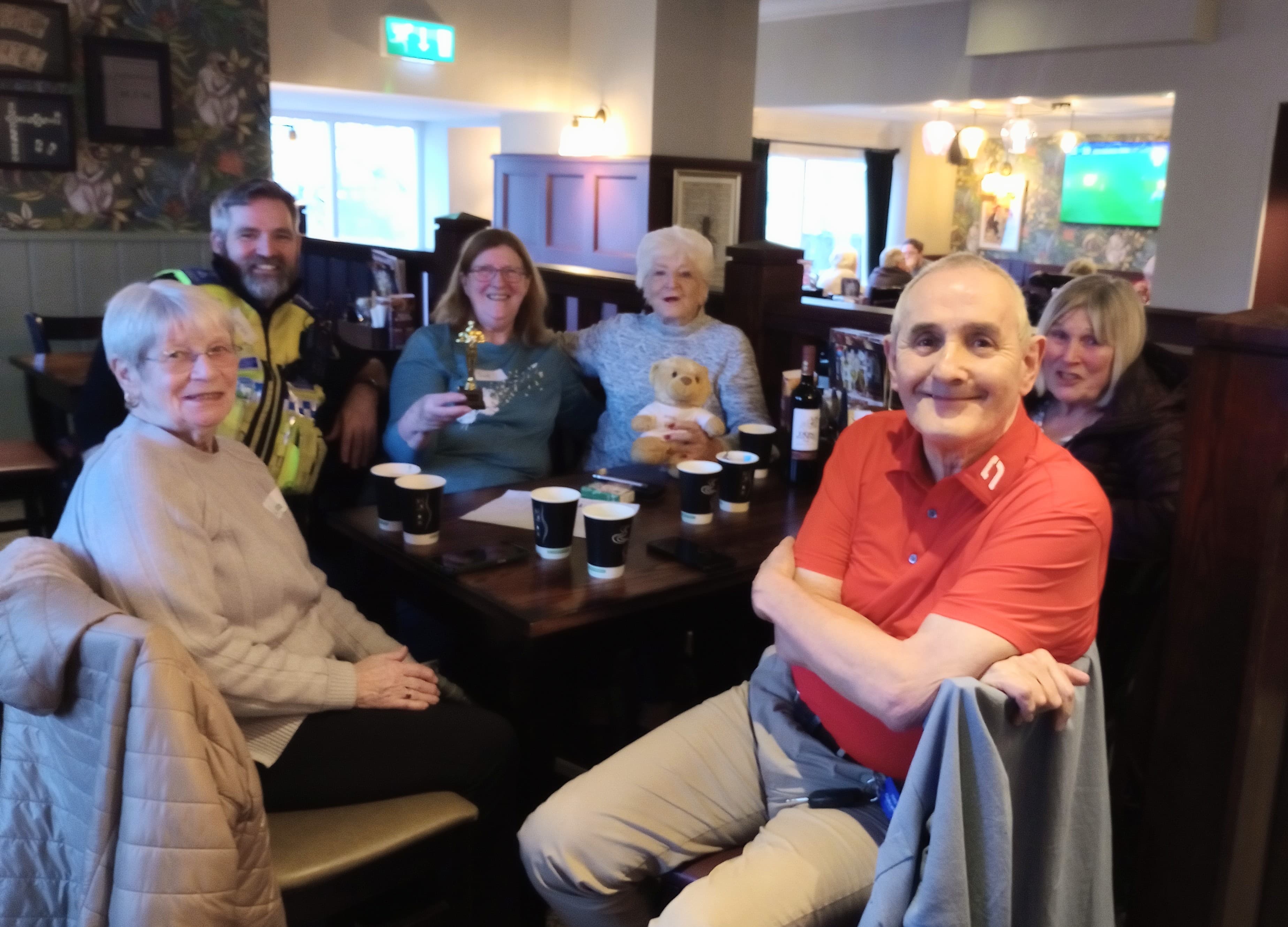 Six smiling people sit around a pub table with coffee cups, a teddy bear, and a trophy. - Home Instead